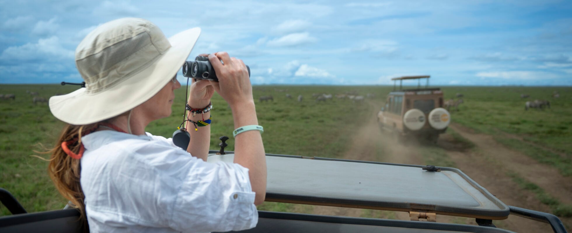 Woman with hat and binoculars standing in a jeep on a road in Tanzania